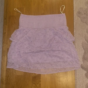 Ljuslila kjol med volang - Söt ljuslila kjol med volangdetaljer och broderade blommor. Kjolen har en elastisk midja för bekväm passform och är perfekt för sommarens varma dagar. Den är kort och har en luftig design.