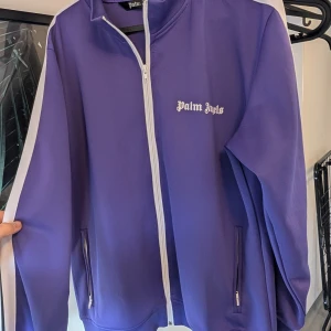 Lila jacka från Palm Angels - Snygg lila jacka från Palm Angels med vit dragkedja och logga på bröstet. Jackan har en sportig look med långa ärmar och ribbade detaljer vid kragen och ärmsluten. Perfekt för en stilren och avslappnad stil.