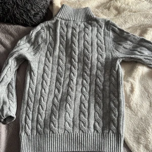 Grå stickad tröja från Ralph Lauren - Säljer en stilren grå stickad tröja från Ralph Lauren med kabelstickat mönster och dragkedja vid halsen. Tröjan har en hög krage och långa ärmar, perfekt för kyliga dagar. En klassisk och tidlös design som passar till mycket.