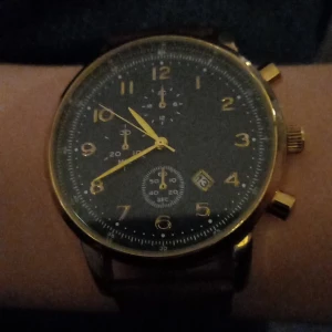 Byter mot en moncler  - Snygg herrklocka från Kiomi med brun läderrem och guldiga detaljer. Urtavlan är svart med guldiga siffror och visare, samt har en datumvisare. Perfekt för den stilmedvetne!