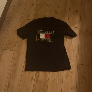 Svart t-shirt från Tommy Hilfiger - Snygg svart t-shirt från Tommy Hilfiger med ett stort tryck på framsidan i guld, vitt och rött. Perfekt för en casual look med en touch av stil. Passar bra till jeans eller shorts.