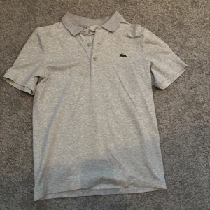 Grå pikétröja från Lacoste - Säljer en stilren grå pikétröja från Lacoste. Tröjan har en klassisk krage och korta ärmar, perfekt för en avslappnad look. Den ikoniska krokodilloggan pryder bröstet. Passar utmärkt till både jeans och shorts.