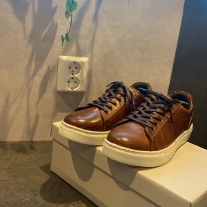 Bruna sneakers i skinn - Snygga bruna sneakers i skinn med vita sulor och svarta snören. Perfekta för en stilren look. Skorna har en klassisk design med rund tå och snörning framtill.