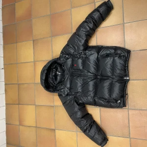 Polo Ralph lauren jacka - Säljer en svart pufferjacka med huva och dragkedja. Jackan har långa ärmar och är perfekt för kalla dagar. Den är tillverkad i ett glansigt syntetmaterial och har en praktisk design med flera fickor.