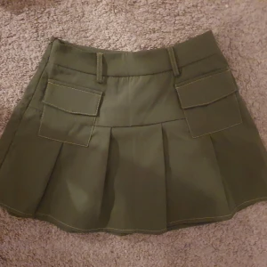 Olivgrön kortkjol med fickor - Snygg olivgrön kortkjol med små fickor framtill. Kjolen har en stilren design med veckade detaljer som ger en fin form. Perfekt för en avslappnad och trendig look.