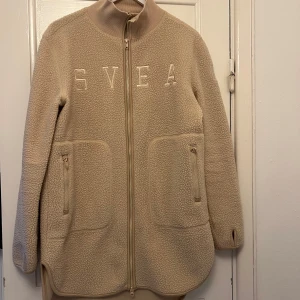 Beige fleecejacka från Svea - Mysig beige fleecejacka från Svea med dragkedja och broderad logga på bröstet. Jackan har två praktiska fickor med dragkedjor framtill och en hög krage för extra värme. Perfekt för kyliga dagar!