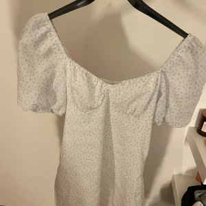 Sommarklänning - Söt vit klänning med blå blommor och puffiga kortärmar. Perfekt för sommarens varma dagar. Klänningen har en smickrande passform och en romantisk känsla.