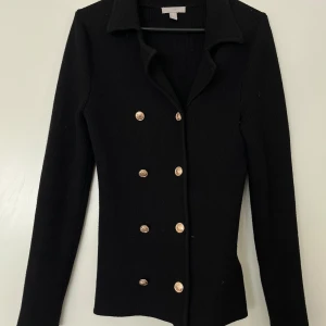 H&M svart kavaj/kofta XS - Svart, figursydd kavaj/kofta med dubbelknäppning och guldiga knappar. Elegant modell som passar både till vardags och fest. Köpt från H&M. Storlek XS. I fint skick. 46% viskos, 29% polyester och 25% polyamide.   Mått: Längd ca 70 cm Byst ca 32 cm Midja ca 32 cm Ärmlängd mätt från axeln ca 65 cm Axelmått ca 40 cm