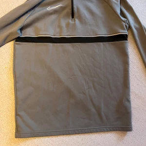 Grå tröja från Nike - Säljer en grå tröja från Nike med dragkedja och Dri-FIT teknologi. Tröjan har långa ärmar och en stilren design med en vit Nike-logga på bröstet. Perfekt för träning eller vardagsbruk.