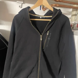 Svart hoodie från Sail Racing - Säljer en svart hoodie från Sail Racing med dragkedja och broderad logga på bröstet. Jackan har en huva och en praktisk ficka med dragkedja. Perfekt för en casual look.