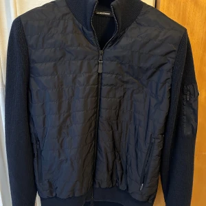 Marinblå jacka från J.Lindeberg - Snygg svart jacka från J.Lindeberg med quiltad framsida och stickade ärmar. Jackan har dragkedja framtill och en diskret logotyp på ärmen. Perfekt för en stilren look. Pris kan diskuteras vid snabb affär.