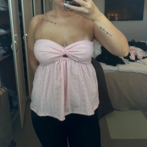 Rosa axelbandslös topp - Säljer en söt rosa axelbandslös topp med knytdetalj framtill. Toppen har ett fint hålmönster och är perfekt för varma dagar. Passar bra till både jeans och kjol.