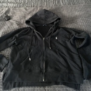 Svart hoodie från Ralph Lauren - Säljer en klassisk svart hoodie från Ralph Lauren med dragkedja och huva. Den har en liten logga på bröstet och snören vid huvan. Perfekt för en avslappnad stil.