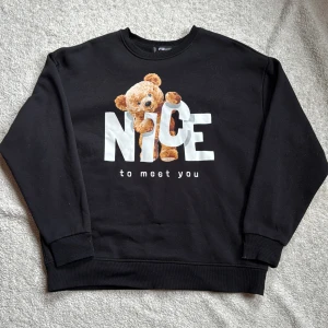 Svart sweatshirt med nallemotiv från FB Sister - Säljer en svart sweatshirt från FB Sister med ett gulligt nallemotiv och texten 'Nice to meet you' på framsidan. Tröjan har långa ärmar och en bekväm passform. Perfekt för en avslappnad stil!