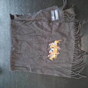 Svart halsduk från Polo Ralph Lauren - Snygg svart halsduk från Polo Ralph Lauren med broderat motiv av polospelare i färgglada kläder. Halsduken har fransar i ändarna och är tillverkad av mjuk ull för extra värme. Perfekt för kyliga dagar! 100% Lamm-ull (Väldigt fint material) Nypris 999 kr Vårt pris 249 kr