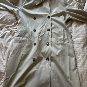 Beige trenchcoat med knappar - Snygg beige trenchcoat med dubbelknäppning och klassisk krage. Perfekt för vår och höst. Jackan har långa ärmar och två fickor framtill. En tidlös klassiker för garderoben.