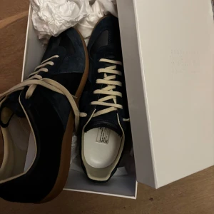 Maison Margiela skor - Snygga marinblå sneakers från Maison Margiela med mockadetaljer och vita snören. Skorna har en klassisk design med en gummisula i brunt. Perfekta för en stilren look.