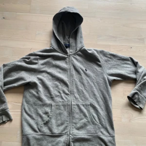 Grå hoodie från Polo Ralph Lauren - Säljer en stilren grå hoodie från Polo Ralph Lauren med dragkedja och broderad logga på bröstet. Tröjan har långa ärmar och en praktisk huva. Perfekt för en avslappnad look.