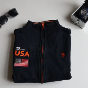  U.S. Polo Assn. Cardigan  -  svart fleecejacka från U.S. Polo Assn. med orange detaljer och USA-flagga. Jackan har dragkedja och broderad logga på bröstet. Original pris var 889 men säljer den billigare.