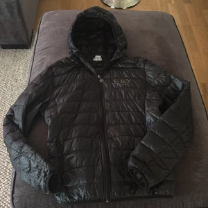 Svart pufferjacka från Emporio Armani - Snygg svart pufferjacka från Emporio Armani med huva och dragkedja. Jackan har ett stilrent quiltat mönster och EA7-logga på bröstet. Perfekt för kyligare dagar.