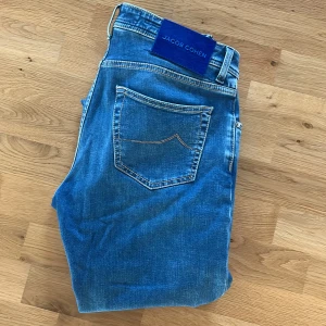 Blå jeans från Jacob Cohen - Snygga blå jeans från Jacob Cohen med klassisk femficksdesign och kontrastsömmar. Perfekta för en stilren look. Märkesdetaljer på baksidan och insidan av midjan. Nypriset ligger på 5500 de är aldrig använda  och har inga skador. Modellen är nick och i storlek 34 men passar storlek 32-33