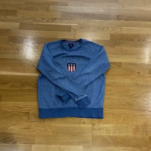 Blå sweatshirt från GANT - Säljer en snygg blå sweatshirt från GANT med ett broderat emblem på bröstet. Tröjan har långa ärmar och rund halsringning. Perfekt för en avslappnad stil.