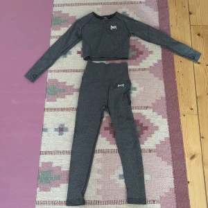 Grå croppad träningstopp och leggings med scrunch - Säljer ett set med grå croppad träningstopp och matchande leggings. Toppen har långa ärmar och en stilren design med en liten logga på bröstet. Leggingsen har hög midja och en logga på höften. Perfekt för träning eller yoga! Står storlek s men skulle säga att det är xxs-xs