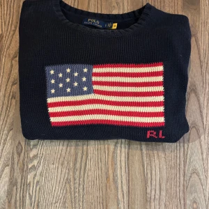 Polo Ralph Lauren med amerikanska flaggan  - Säljer en snygg mörkblå tröja från Polo Ralph Lauren med en stor amerikansk flagga på framsidan. Tröjan är tillverkad i 100% bomull och har en klassisk rund halsringning. Perfekt för en stilren look! Ny pris ligger på 6000. Den är köpt i en av deras butiker