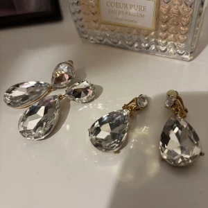 Clips örhängen med droppformade stenar - Säljer 2 par eleganta clipsörhängen med stora, glittriga droppformade stenar. Passar perfekt till både bal, bröllop eller andra lyxiga tillfällen. Örhängena är från Balunz och Åhléns och är i nyskick.