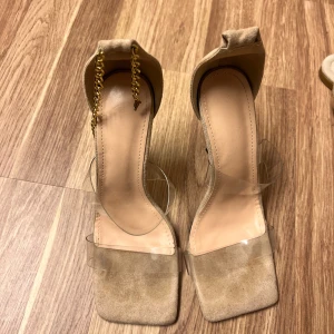 Beige klackskor med kedjedetalj - Eleganta beige klackskor med transparent rem och kedjedetalj vid hälen. Skorna har en fyrkantig tå och är tillverkade i mocka. Perfekta för en stilren look.