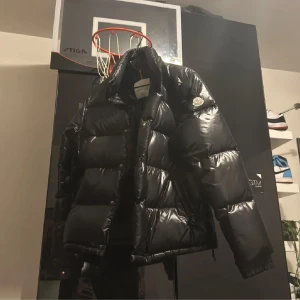 Svart pufferjacka från Moncler - Säljer en snygg svart pufferjacka från Moncler. Jackan har en glansig finish och är perfekt för kyligare väder. Den har långa ärmar och en klassisk Moncler-logga på ärmen. Perfekt för dig som vill hålla stilen i vinter! OBS cartoon stitching saknas och en knapp för att göra armarna tightare