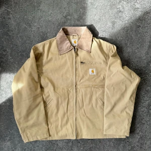 Beige jacka från Carhartt - Säljer en beige jacka från Carhartt med dragkedja och en praktisk bröstficka. Jackan har en kontrasterande krage i manchester och är perfekt för både höst och vår. Den har en klassisk design med Carhartt-logga på bröstet. Storlek  L o cropped fit Reworked 