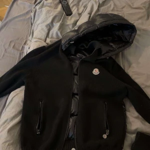 Svart jacka från Moncler - Säljer en snygg svart jacka från Moncler med huva och dragkedja. Jackan har en quiltad insida och stickad utsida, vilket ger en cool kontrast. Perfekt för kyligare dagar!