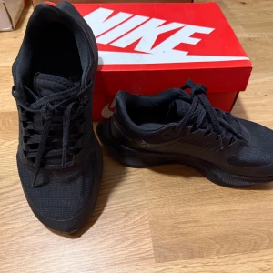 Svarta Nike Pegasus 38 Shiled löparskor - Snygga svarta Nike löparskor med Nike Shield-teknologi för extra skydd mot väder. Skorna har en stilren design med snörning och en diskret logga på sidan. Perfekta för löpning och träning.