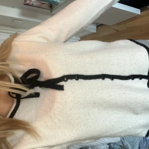 Vit tröja med svarta detaljer från Nelly - Säljer en stilren vit tröja från Nelly med svarta detaljer. Tröjan har en dekorativ svart rosett vid halsen och knappar framtill. Perfekt för en chic look!