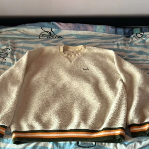 Vit fleece tröja från Levi's i M - Säljer en vit fleece tröja från Levi's med broderad logga på bröstet. Tröjan har en avslappnad passform och ribbade muddar med ränder i orange och grön vid ärmslut och nederkant. Perfekt för kyliga dagar!