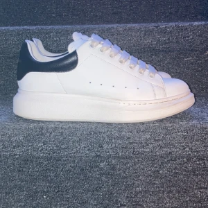 Alexander McQueen sneakers - Snygga vita sneakers från Alexander McQueen med svart häl och klassisk snörning. Skorna har en chunky sula som ger extra höjd och stil. Perfekta för att lyfta vilken outfit som helst. Skick: 8/10 pågrund av under sulan och små smuts som inte går bort men som inte alls är så synligt. Man får med själva boxen och påsen  dessutom i köpet!