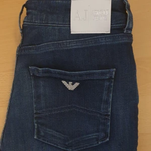 Mörkblå jeans från Armani Jeans - Säljer ett par äkta mörkblå jeans från Armani Jeans med klassisk femficksdesign och silverfärgad logga på bakfickan. Jeansen har dragkedja och knapp framtill samt normal passform. Perfekta till vardags för en stilren look. Skinny passform så hade passat utmärkt för att styla med boots, klippa av till shorts eller bara ha som de är! Midjemått rakt över 35 cm. Innerbenslängd 63 cm.
