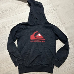 Mörkblå hoodie från Quiksilver - Säljer en mörkgblå hoodie från Quiksilver med röd logga på bröstet. Tröjan har huva och en stor magficka framtill. Perfekt för en avslappnad stil och enkel att matcha med jeans eller joggers. Fint använt skick. 