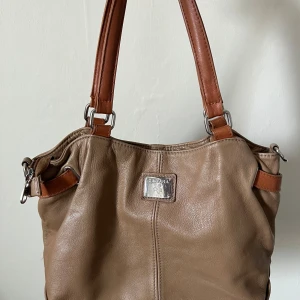 Beige handväska från Zagara  - Snygg, vintage handväska från Zagara i skinn med bruna handtag och silverfärgade metalldetaljer. Väskan har flera fack med dragkedja både utvändigt och invändigt, perfekt för att organisera dina saker. Köp secondhand, säljer då den inte används längre.
