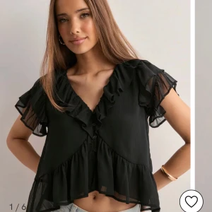 Svart blus med volang och v-ringning - Snygg svart blus med v-ringning och volangdetaljer längs halsringningen och nederkanten. Blusen har korta, transparenta ärmar och knappar framtill. Perfekt för en trendig och feminin look. Köpt i helgen och aldrig använd! 