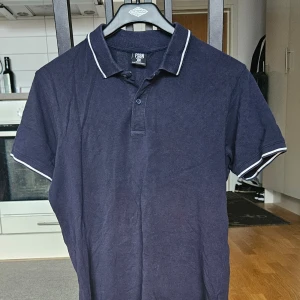 Mörkblå pikétröja från FSBN - Säljer en mörkblå pikétröja från FSBN i slim fit-modell. Tröjan har korta ärmar och vit kant på krage och ärmslut. Perfekt för en stilren och sportig look.