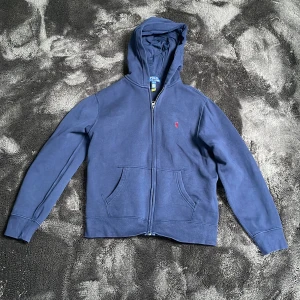 Blå hoodie från Polo Ralph Lauren - Säljer en klassisk blå hoodie från Polo Ralph Lauren med dragkedja och huva. Tröjan har två fickor framtill och den ikoniska lilla röda loggan broderad på bröstet. Perfekt för en avslappnad stil. Storlek 158/164.