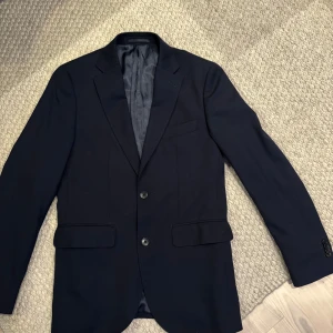 Navy kavaj från Riley - Säljer en klassisk Navy kavaj från Riley med två knappar framtill och fickor med lock. Perfekt för bal eller fest. Kavajen har en stilren design och är helfodrad för extra komfort.