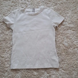 Vit ribbad t-shirt från Lindex - Säljer en enkel och stilren vit ribbad t-shirt från Lindex. T-shirten har korta ärmar och rund halsringning, perfekt för en clean och fräsch look. Passar dig som gillar basic plagg med en twist.