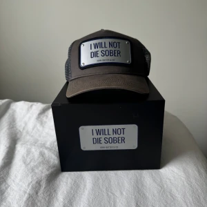 Grå metall keps från John Hatter & Co - Cool grå keps med justerbar baksida och meshpaneler. Framtill sitter en metallskylt med texten 'I WILL NOT DIE SOBER'. Perfekt statement-piece för dig som vill sticka ut. Hör av er vid funderingar ✅