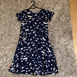 Mörkblå klänning med vitt fjärilsmönster - Mörkblå klänning med korta ärmar och vitt fjärilsmönster över hela plagget. Klänningen har rund halsringning och en utställd kjol som ger en fin siluett. Perfekt för dig som gillar lekfulla mönster och bekväm passform.