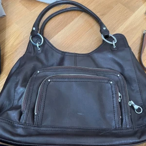 Mörkbrun handväska från Franco Sarto - Säljer en mörkbrun handväska från Franco Sarto i skinn med silverfärgade metalldetaljer. Väskan har dubbla handtag, en stor ytterficka med dragkedja och ett färgglatt prickigt innerfoder. Perfekt för dig som vill ha en stilren och rymlig accessoar.