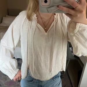 Vit blus med spetsdetaljer - Säljer en luftig vit blus med v-ringning och knytband framtill. Blusen har långa ärmar med resår vid ärmslut och fina spetsdetaljer längs sidorna. Perfekt till jeans för en avslappnad men stilren look.