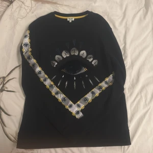 Svart sweatshirt med ögonmönster från Kenzo - Säljer en svart sweatshirt från Kenzo i storlek L. Tröjan har ett stort ögonmönster framtill i vitt, blått och gult samt matchande detaljer längs ärmarna. Rund halsringning med gul kant. Perfekt statement-plagg för dig som gillar unika prints.